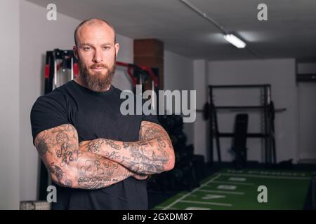 Sportif adulte fort avec barbe et tatouages sur les bras pliés regardant la caméra dans le gymnase Banque D'Images
