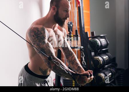 Vue latérale de la récolte sportif musclé concentré avec tatouages faire des exercices sur la machine de câble croisé dans la salle de gym avec des murs légers Banque D'Images