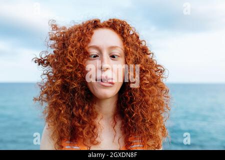 Une femelle enjouée aux cheveux bouclés montrant la langue et croisant les yeux tout en faisant de la grimace drôle sur la côte d'une mer bleue ondulée Banque D'Images