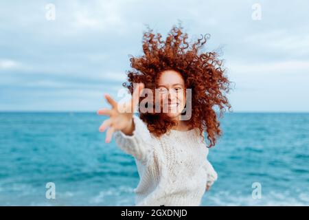 Agréable femme avec long gingembre curly hairstyle atteignant la main à l'appareil photo tout en se tenant contre la mer agitant Banque D'Images