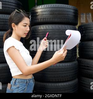 Le jeune propriétaire asiatique du magasin de pièces automobiles vérifie le nombre de pneus et les commandes reçues avant de commencer le travail le matin. Plusieurs pneus de voiture ar Banque D'Images