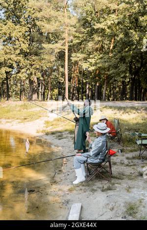 Des hommes multiculturels âgés pêchant dans le lac près d'un ami excité dans le parc Banque D'Images