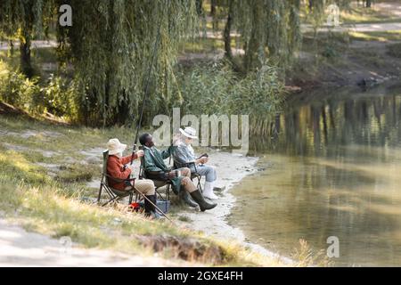 Hommes multiethniques âgés avec canne à pêche assis près du lac dans le parc Banque D'Images