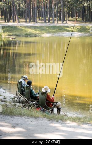 Amis multiethniques âgés avec canne à pêche assis près du lac dans le parc Banque D'Images