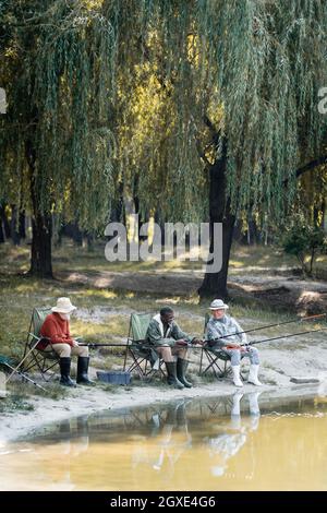 Des hommes multiculturels et gais en bottes de caoutchouc pêchant sur des chaises près du lac dans le parc Banque D'Images