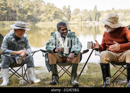 Souriant hommes multiethniques dans une tenue de pêche tenant des thermo-tasses tout en parlant sur des chaises près du lac Banque D'Images