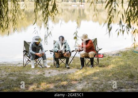 Homme senior avec coupe thermo discutant avec des amis interraciaux en tenue de pêche sur la côte du lac Banque D'Images