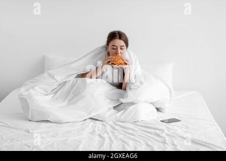 Une jeune femme triste et affamée est assise sur un lit blanc à la maison et mange du hamburger, souffrant de dépression et de problèmes mentaux Banque D'Images