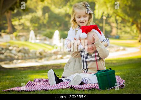 Petite fille douce essaie de mettre un chapeau de Père Noël sur son petit frère réticent à l'extérieur dans le parc. Banque D'Images