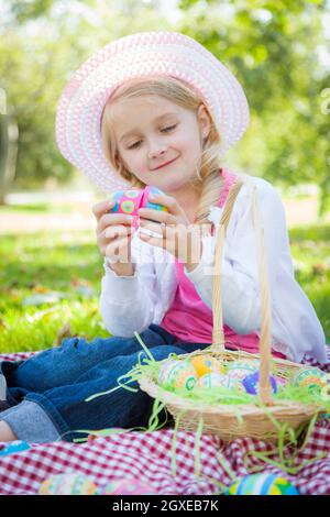 Mignon jeune fille sur pique-nique couverture portant chapeau aime ses oeufs de Pâques dehors dans le parc. Banque D'Images