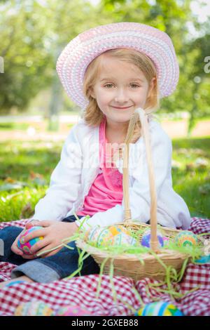 Mignon jeune fille sur pique-nique couverture portant chapeau aime ses oeufs de Pâques dehors dans le parc. Banque D'Images