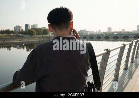 Vue arrière d'un jeune homme d'affaires avec un téléphone intelligent qui vous appelle au bord de la rivière Banque D'Images
