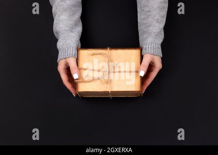 Femme mains en tricot gris chandail avec ongles blancs tenant en bois rustique boîte cadeau décorée avec corde de chanvre sur fond noir. Vue de dessus, plan de travail c Banque D'Images