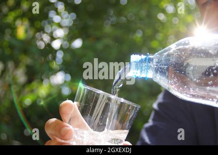 Eau versée dans un verre à la main avec le jardin en arrière-plan. Rafraîchissement avec de l'eau minérale propre naturelle dans une bouteille en plastique. Banque D'Images