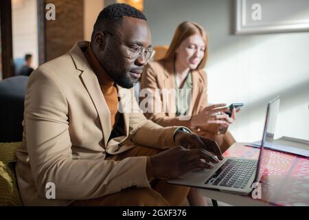Jeune homme africain sérieux en lunettes et en tenue de formalwear regardant l'écran d'ordinateur portable contre une collègue féminine Banque D'Images
