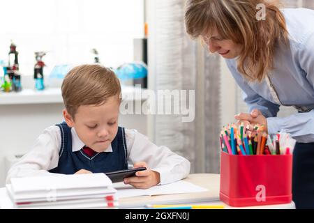 Un jeune garçon de première classe mignon dans un uniforme d'école à la maison joue sur un smartphone à un bureau avec des livres et des crayons. Maman le grimpa. Mise au point sélective. Gros plan. Banque D'Images