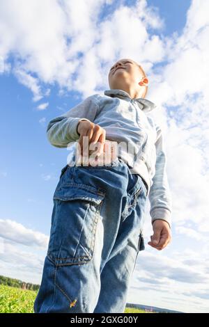 Un enfant d'âge préscolaire satisfait en jeans et un chandail tient l'argent de papier dans la nature dans un champ sur le fond d'un ciel bleu avec des nuages. Portrait Banque D'Images