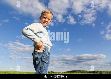 Un enfant d'âge préscolaire satisfait en jeans et un chandail tient l'argent de papier dans la nature dans un champ sur le fond d'un ciel bleu avec des nuages. Portrait Banque D'Images