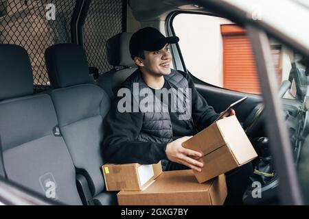 Un messager mâle assis sur le siège du conducteur avec des boîtes en carton et une tablette numérique qui regarde loin Banque D'Images