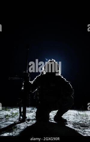 Tireur d'élite avec grand fusil assis tenant le fusil sur fond noir. Un tueur solitaire au clair de lune Banque D'Images