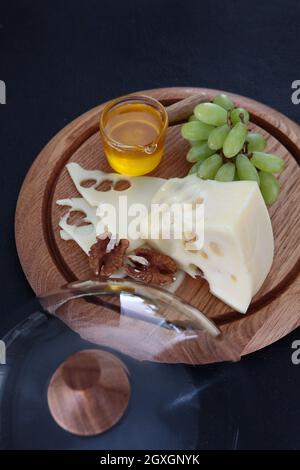 Fromage à pâte dure avec de grands trous sur une planche ronde en bois. Plateau de fromage rond en bois avec couvercle en verre. Miel et raisins avec tranches de fromage. Tranches de fromage. Banque D'Images