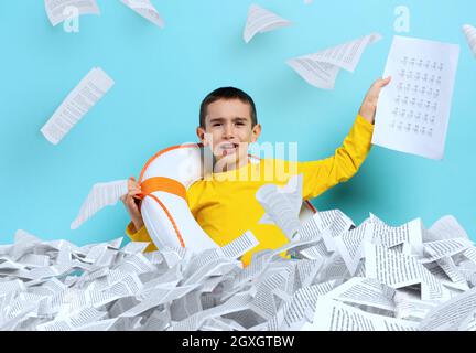 Un jeune garçon malheureux étudiant se noye dans une mer de feuilles d'exercices. Fond cyan Banque D'Images