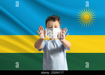 Petit garçon blanc dans un masque protecteur sur le fond du drapeau du Rwanda. Fait un panneau d'arrêt avec ses mains, rester à la maison Rwanda. Banque D'Images