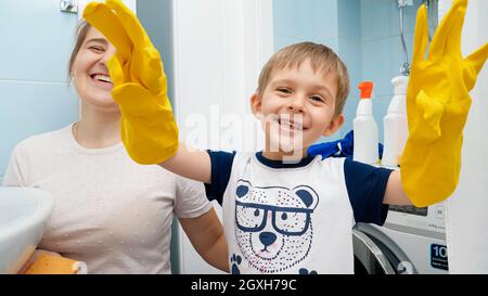 Joyeux sourire petit garçon dans des gants en caoutchouc jaune faisant des travaux ménagers et aidant la mère dans le nettoyage. Banque D'Images