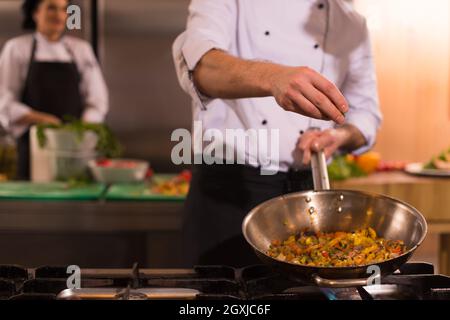 Young male chef mettre les épices sur les légumes dans le wok à cuisine commerciale Banque D'Images