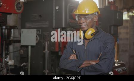 Portrait américain industriel noir jeune homme de travail souriant avec un casque jaune devant la machine, heureux ingénieur debout bras croisés au travail dans le TH Banque D'Images