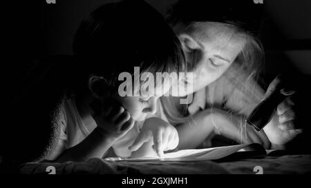 Portrait noir et blanc du livre de lecture de petit garçon avec mère couché sous une couverture la nuit. Banque D'Images