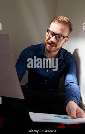 Jeune homme à barbe rouge dans des lunettes assis dans un coin et travaillant avec des papiers financiers dans une pièce sombre Banque D'Images