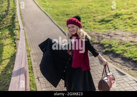 Une jeune fille joyeuse marche dans les escaliers dans une palette bordeaux et le biret, dans des vêtements noirs, dans l'automne sur un fond de nuages bleus. Banque D'Images