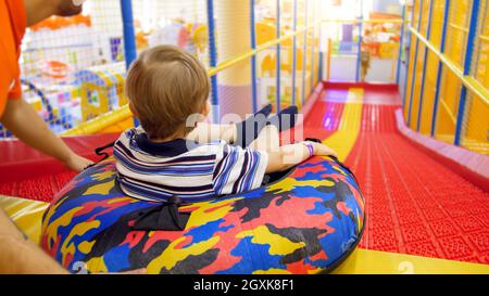 Un petit garçon mignon qui s'enrôle dans le toboggan de l'anneau gonflable en forme de beignet dans la salle de jeux pour enfants du centre commercial. Banque D'Images