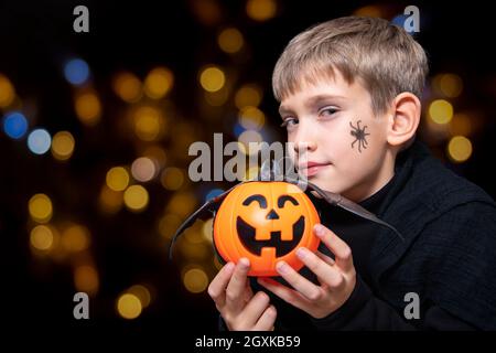 Un enfant tenant un panier orange en forme de citrouille avec un visage de repas, une lanterne de Jack et une chauve-souris sur un fond noir avec bokeh. Le garçon attend pour Banque D'Images