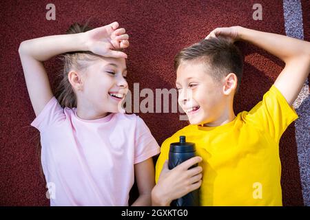 Deux enfants heureux, un garçon et une fille, s'allonger sur le dos et rire en regardant la caméra après un entraînement de jogging sur la piste de course du stade. Sports jumeaux Banque D'Images