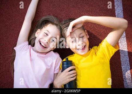 Thème de style de vie de sport pour enfants. Les jeunes garçons et filles heureux s'étendent joyeusement sur la couverture en caoutchouc de la piste de course du stade et s'effacent dans l'appareil photo avec le sourire Banque D'Images