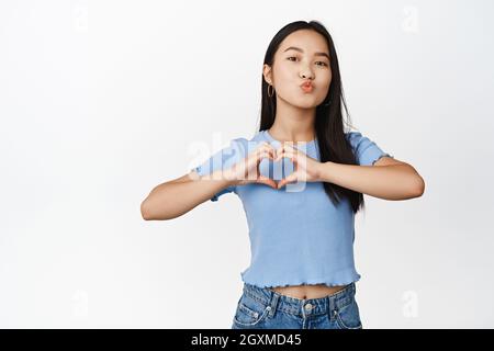 Une femme vietnamienne mignonne montre un signe de coeur et un visage de baiser, debout dans une chemise sur fond blanc Banque D'Images