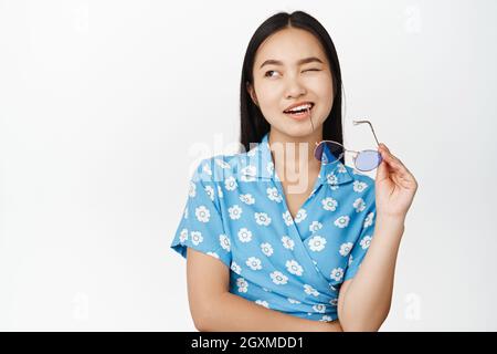 Portrait de la belle fille asiatique moderne bite temple de lunettes de soleil, wencing et sourire avec confiance, debout sur fond blanc Banque D'Images