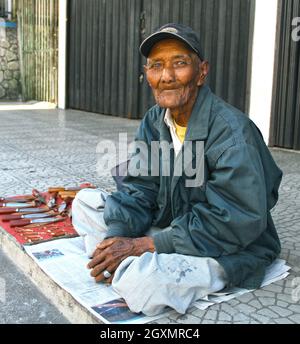 Un vieil homme sans dents assis au bord de la route portant une casquette de baseball vendant des couteaux à Bukittinggi, Sumatra Ouest, Indonésie. Banque D'Images