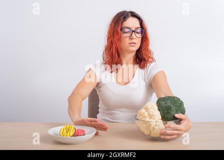 La femme caucasienne préfère une alimentation saine et refuse la restauration rapide. REDHEAD fille choisit entre brocoli et beignes sur fond blanc Banque D'Images