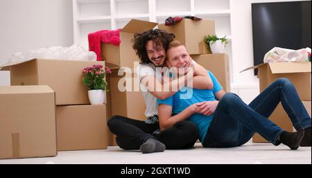 Se détendre dans maison neuve. Cheerful young gay couple assis sur le sol tandis que des boîtes en carton portant tout autour d'eux Banque D'Images