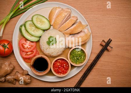 Le délicieux riz au poulet Hainanese est servi avec trois sauces différentes, ainsi que des lamelles de concombre et de tomate dans une assiette sur une table en bois. Vue du dessus Banque D'Images