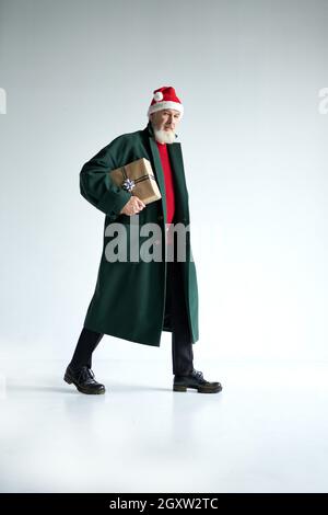 Prise de vue en longueur d'un homme mûr tendance avec chapeau de Noël portant une tenue élégante regardant l'appareil photo, tenant une boîte cadeau tout en marchant isolé Banque D'Images