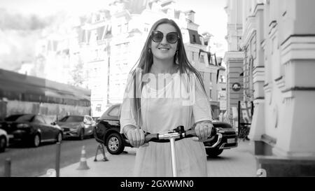 Portrait noir et blanc d'une belle femme souriante dans des lunettes de soleil sur un scooter électrique sur la rue de la ville. Banque D'Images