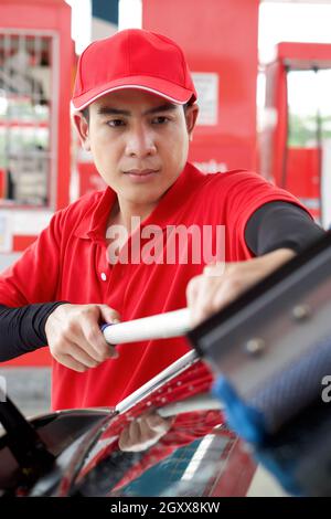 Ouvrier de station-service asiatique en uniforme rouge nettoyant le pare-brise de voiture avec raclette de fenêtre. Banque D'Images
