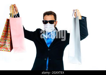 Jeune homme en costume avec masque et lunettes de soleil tenant des sacs de shopping Banque D'Images