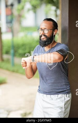 Jeune Indien souriant, avec une barbe debout sur un poteau en bois à l'extérieur et une impulsion de contrôle avec un tracker de forme physique après la course Banque D'Images