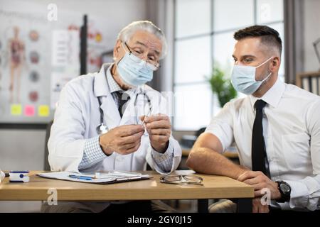 Thérapeute masculin expérimenté montrant à l'homme d'affaires une seringue stérile avec le vaccin de Covid-19.Un patient de sexe masculin se rendant chez un médecin de famille pour recevoir la vaccination. Banque D'Images