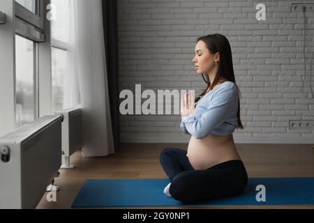 Belle femme enceinte est assise à la maison près de la fenêtre.Elle se replia les mains devant elle et médite. Banque D'Images